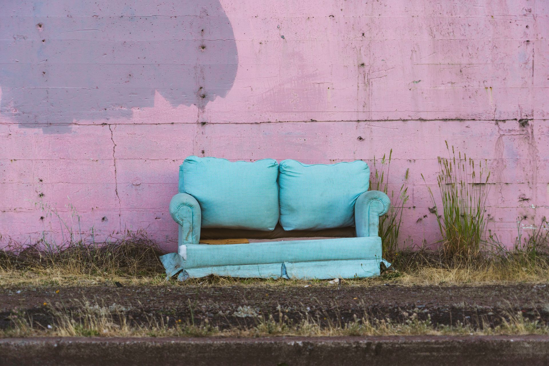 Blue couch in front of a pink, weathered wall. Dry grass and dirt in the foreground.