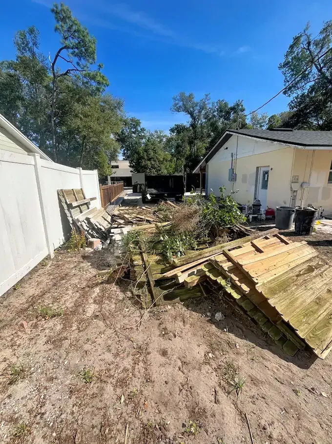 Backyard with debris, including wood and plants. A dumpster sits in the distance, blue sky overhead.