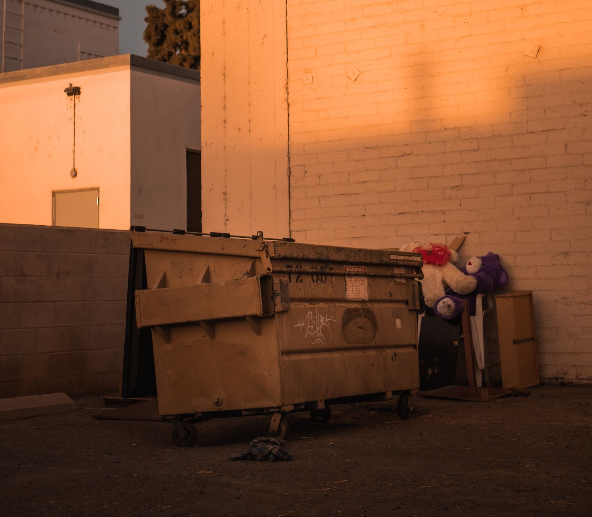 A dumpster in an alley next to a white brick wall and building; toys are leaning against the wall.