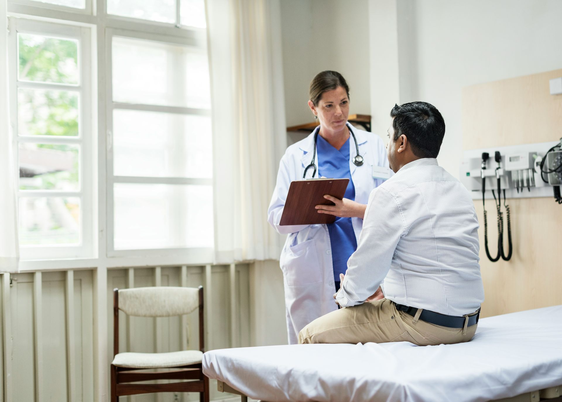 Doctor consulting a seated patient in a bright exam room, holding a clipboard.