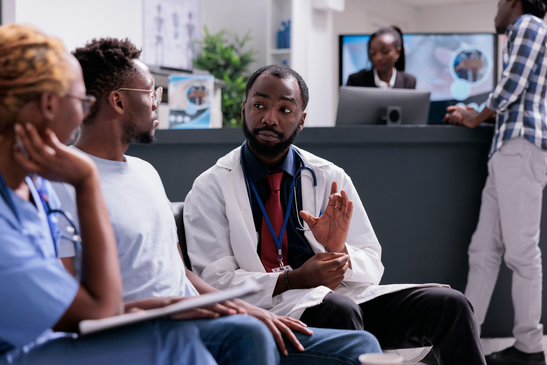 People seated in a clinic waiting area, with a doctor speaking to them and a reception desk in the background.
