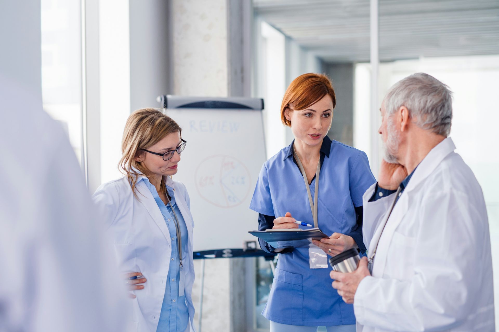 Three medical professionals discussing in a bright hospital hallway, one holding a clipboard.