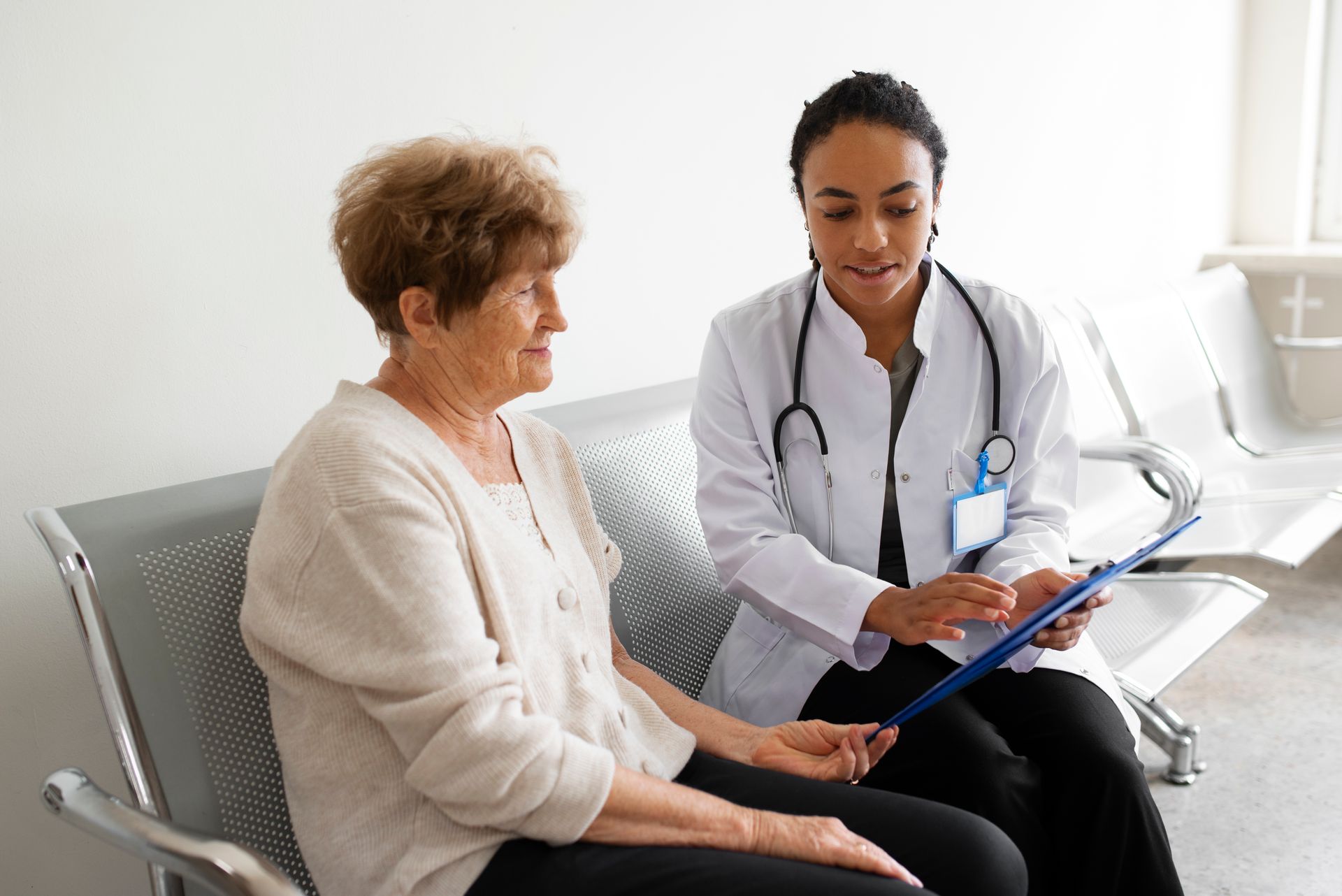 Doctor speaking with hospital patient in bed, holding clipboard in a bright clinic room