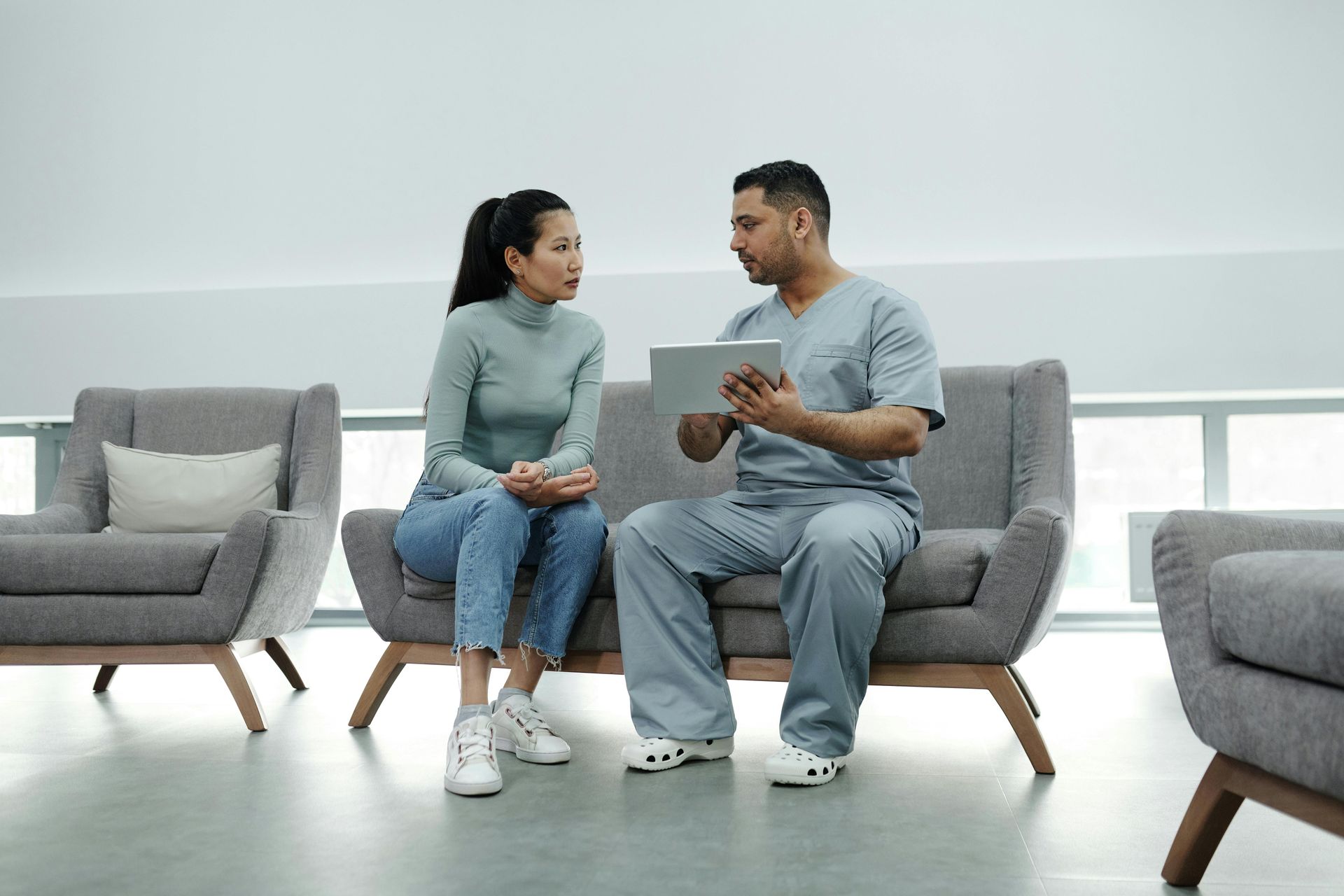 Two people talk on a gray couch in a bright waiting room, holding a clipboard and papers.