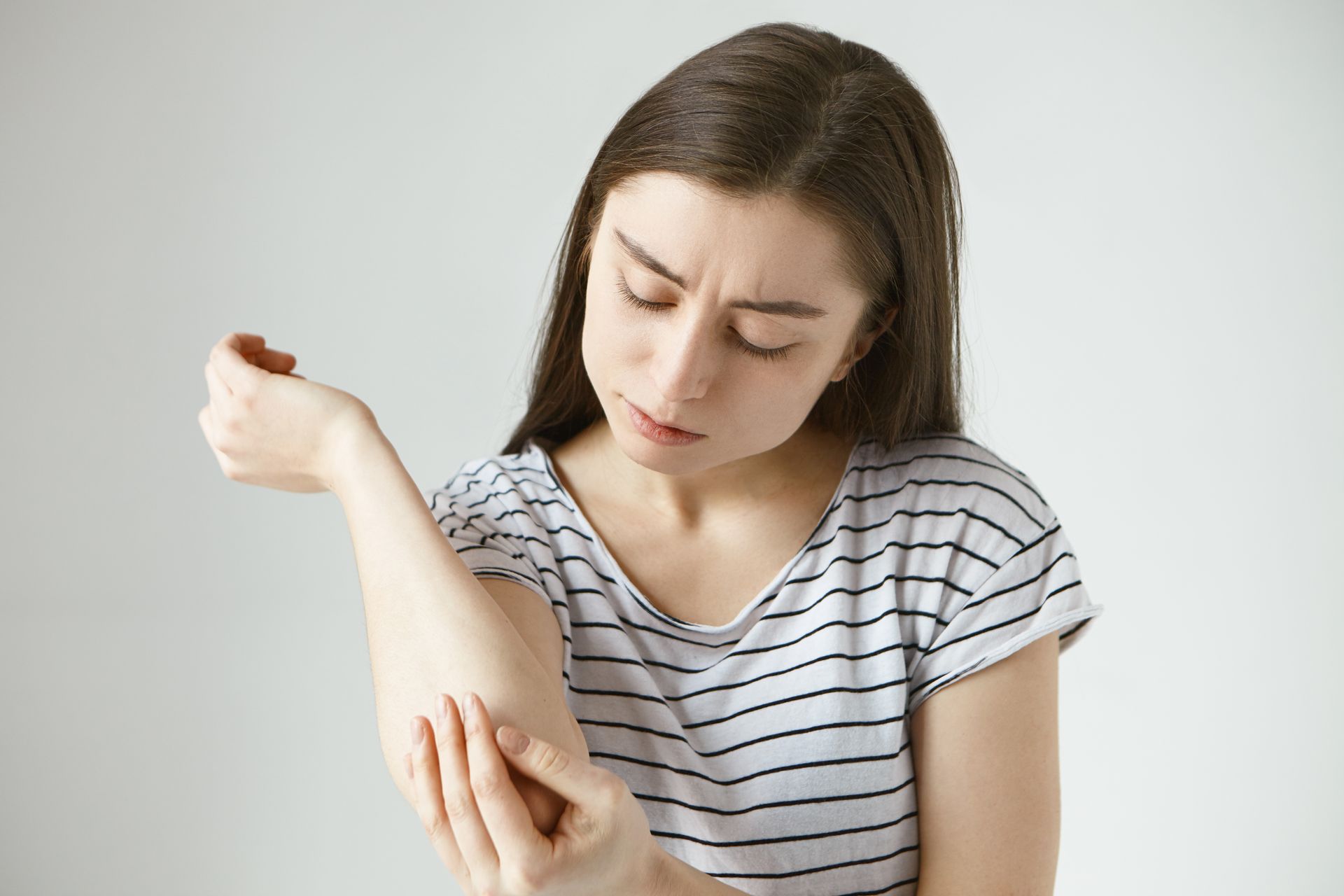 Woman in striped shirt examining an irritated rash on her forearm against a plain background
