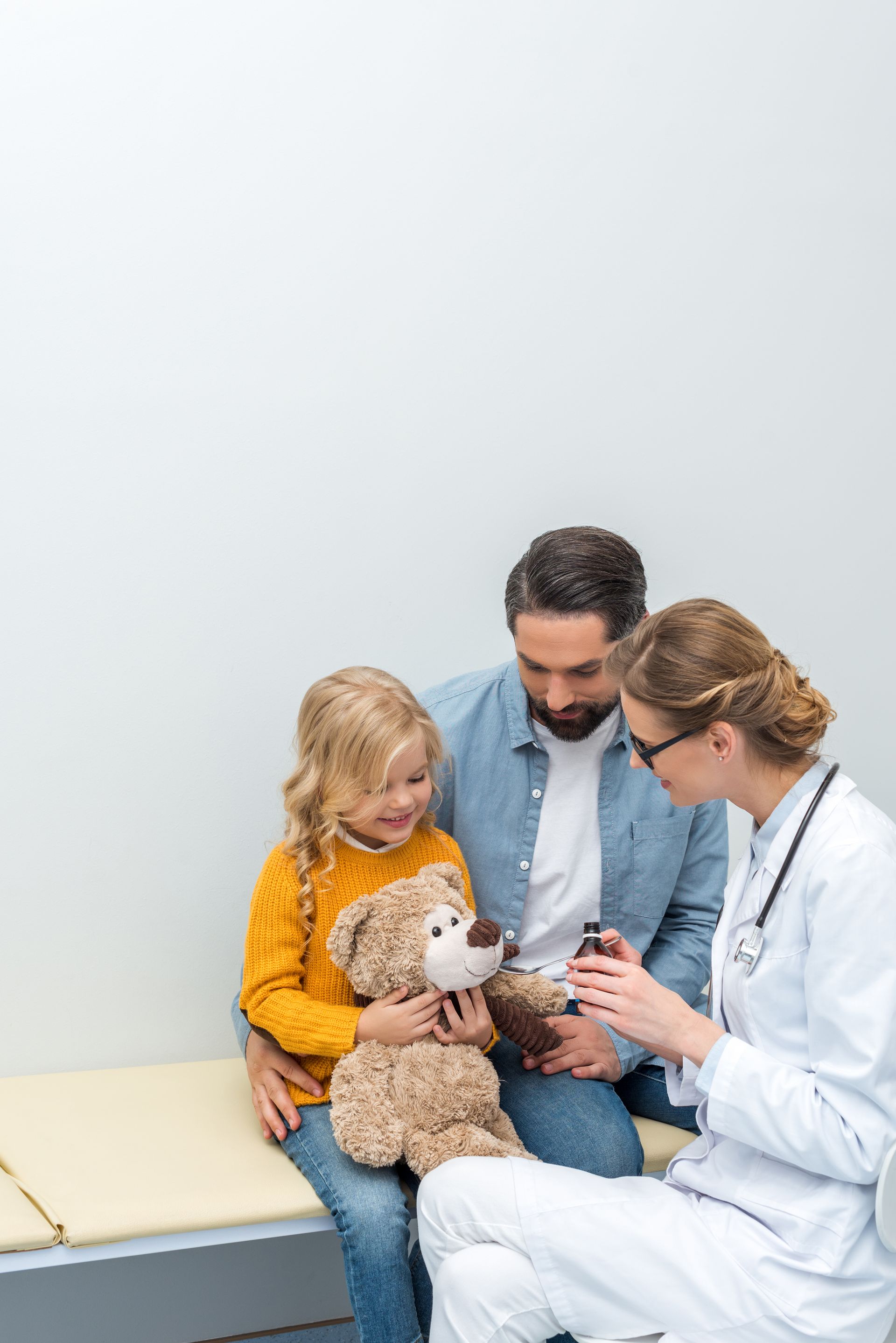 Doctor showing a teddy bear to a child while a parent sits nearby in a clinic.