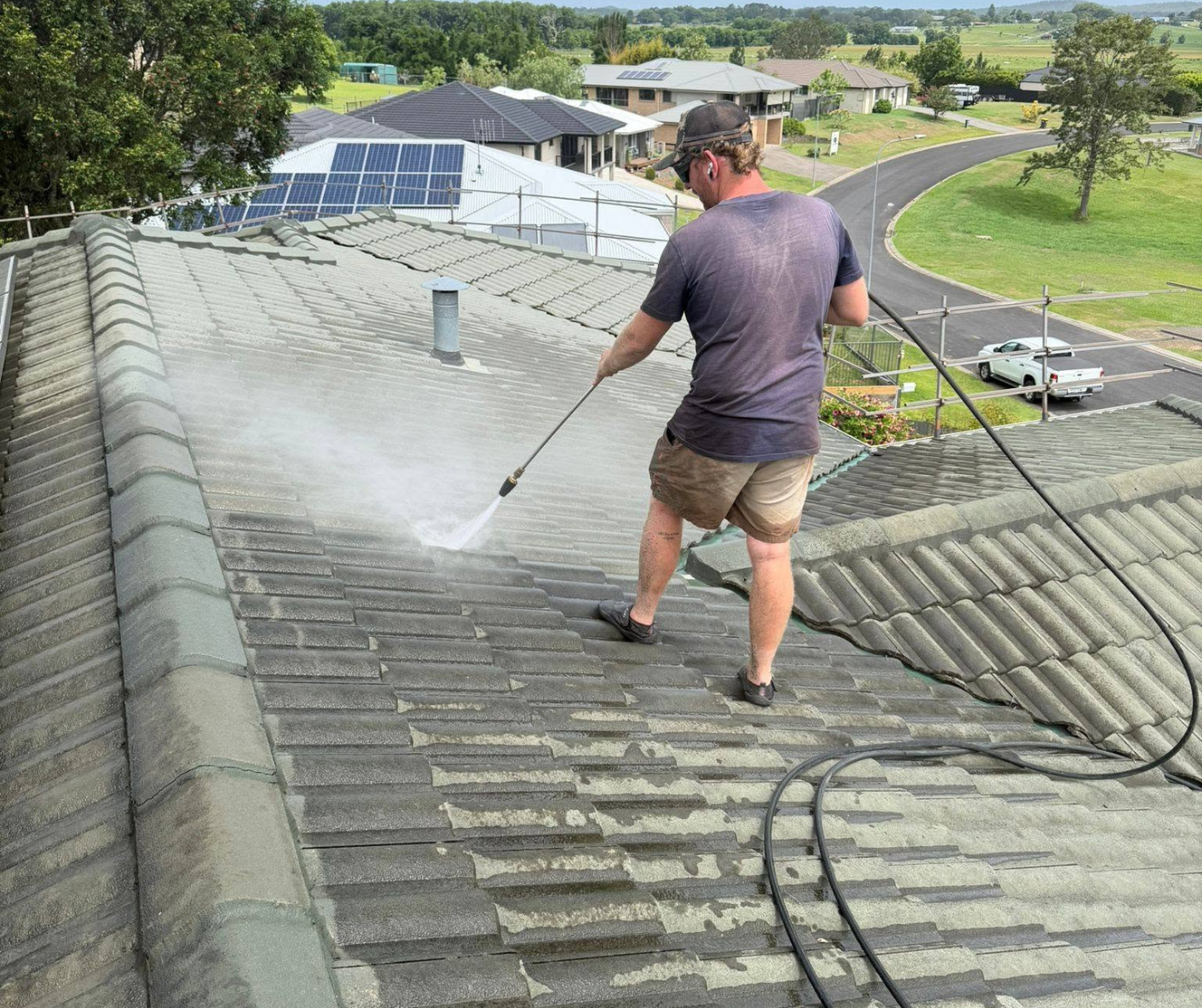 Worker pressure-washing a gray tiled roof on a sunny suburban home