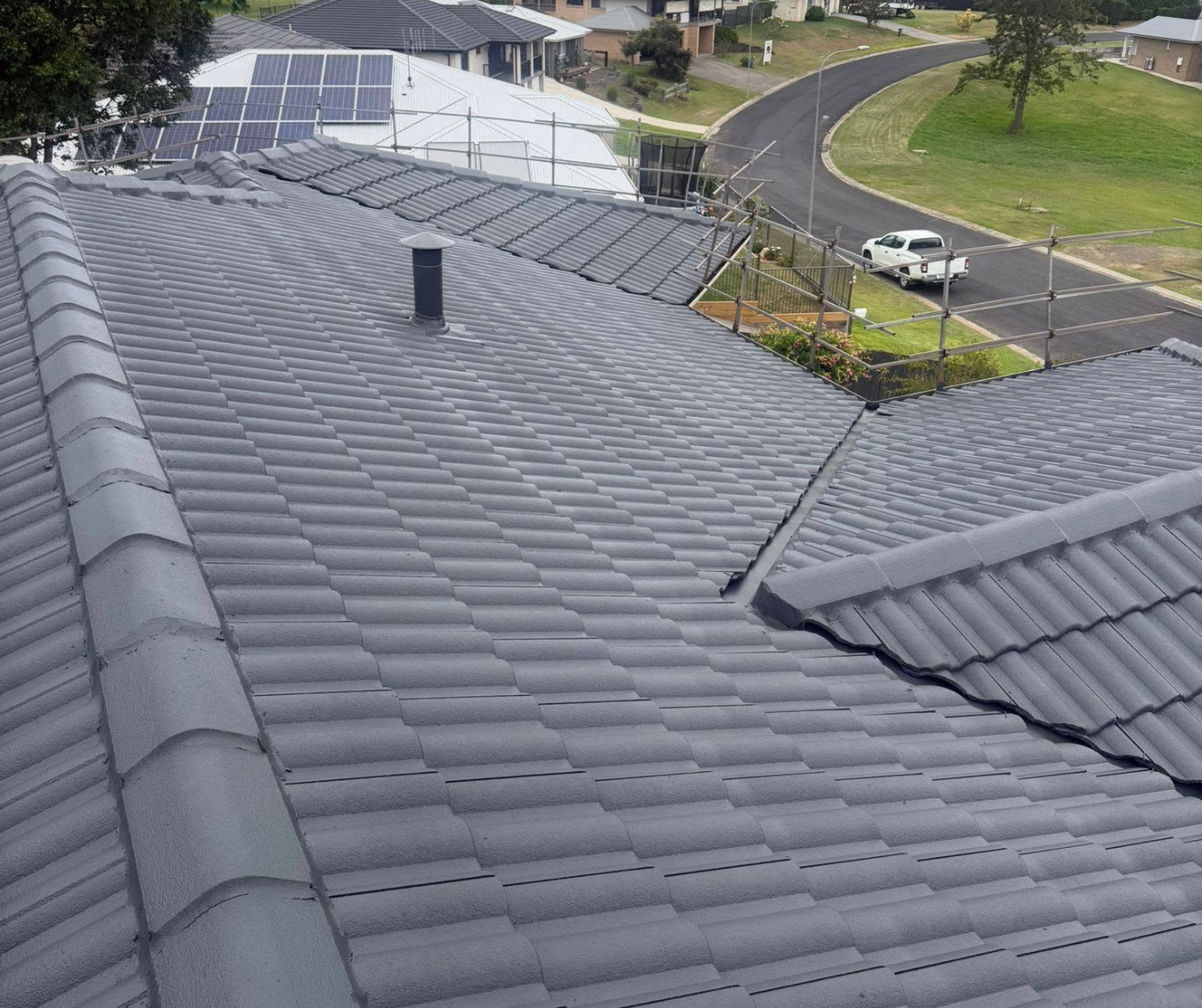 Roof with dark gray tiles, solar panels, and a vent, viewed over a suburban street and houses.