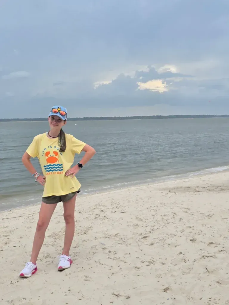 Person standing on a sandy beach in a yellow shirt and pink shoes, with calm water and cloudy sky behind. | Frisco Automotive & Transmission