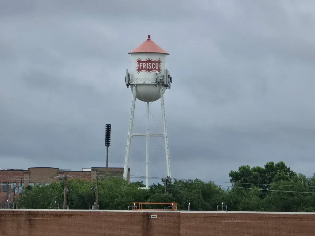 Water tower with a red-roofed top and Hindi text, standing above trees under a cloudy sky. | Frisco Automotive & Transmission