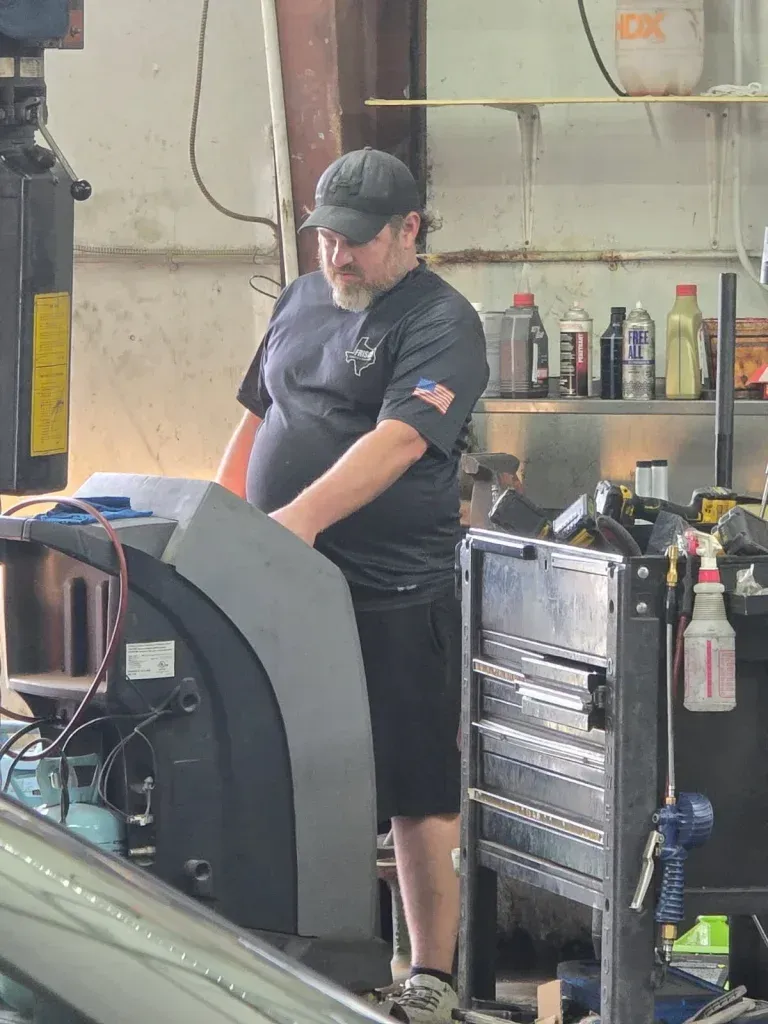 Person in a black cap and shirt standing beside machinery in a workshop. | Frisco Automotive & Transmission