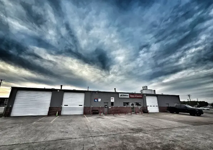 Industrial building with garage doors under a dramatic cloudy sky, with a truck parked on the right. | Frisco Automotive & Transmission
