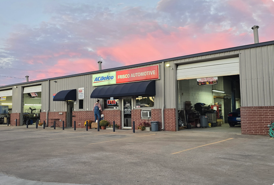 Auto parts and hardware store storefront with open garage bays at dusk | Frisco Automotive & Transmission