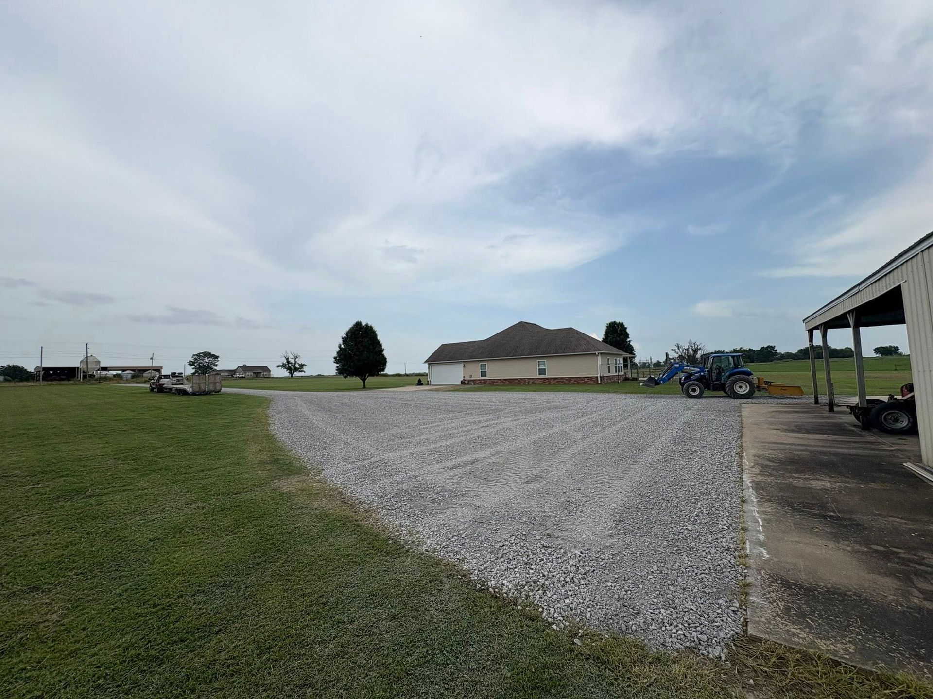 Gravel driveway leading to a house, with a blue tractor, shed, and green field under a cloudy sky.