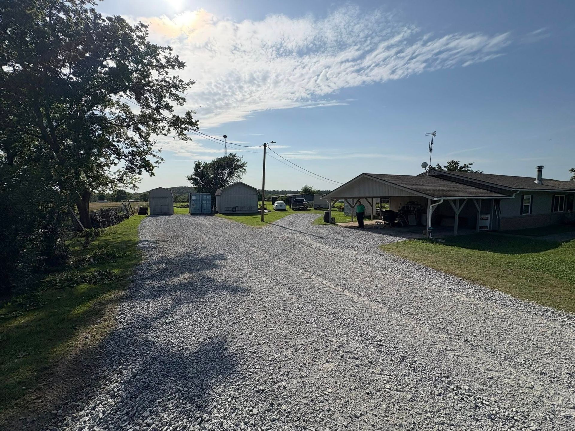 Gravel driveway leads to a house with a carport and outbuildings under a blue sky.
