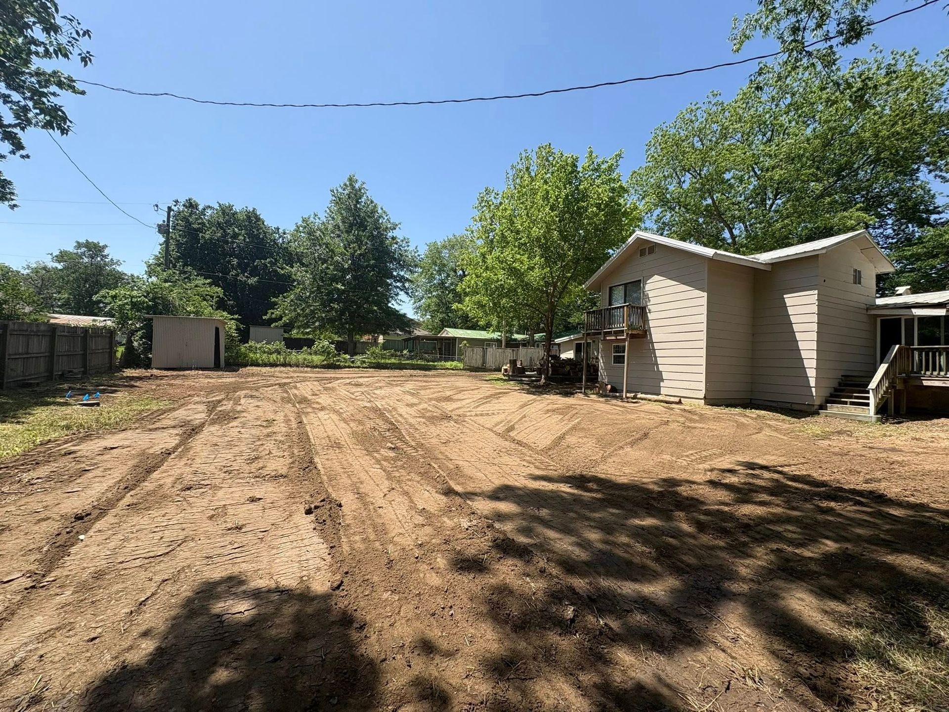Cleared dirt yard next to a light-colored house. Trees and fence in the background, blue sky.