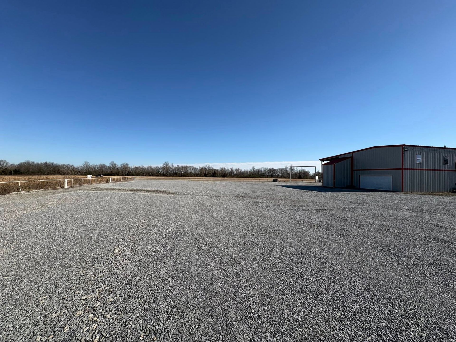 Gravel lot with a metal building on the right, trees in the distance, and a clear blue sky.