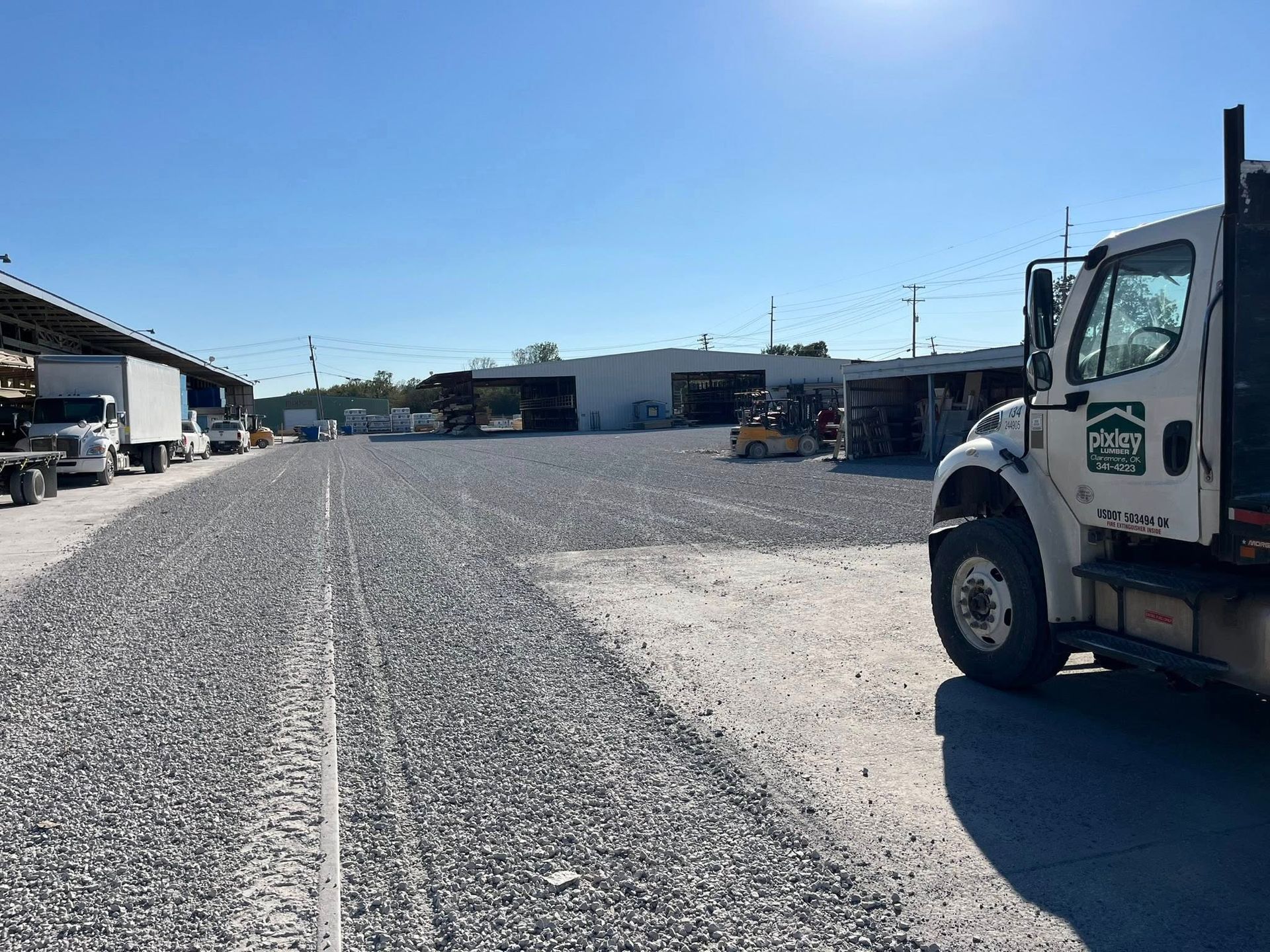 Gravel lot with trucks parked, industrial buildings under blue sky.