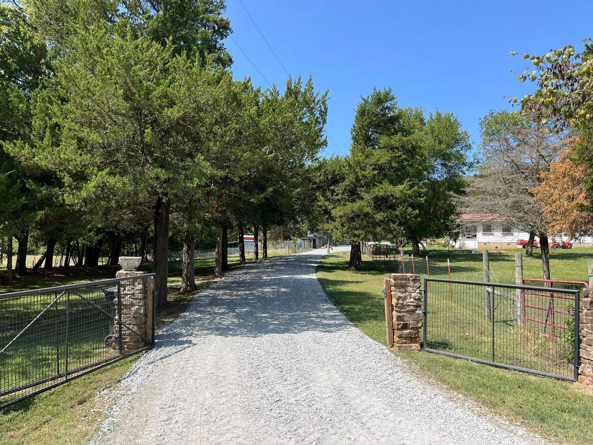 Gravel driveway leading to a building, flanked by trees and a fence on a sunny day.