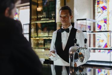 Bartender in black vest and bow tie serves a drink to a patron.
