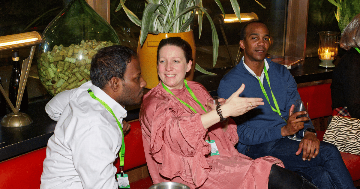 Three people sit on a couch in a dim lounge, smiling and chatting, one gesturing with a hand.