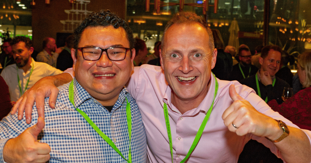 Two smiling men with green lanyards posing together and giving thumbs up at a crowded event