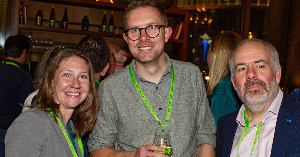 Three people smiling at an indoor event, wearing lanyards and holding drinks.
