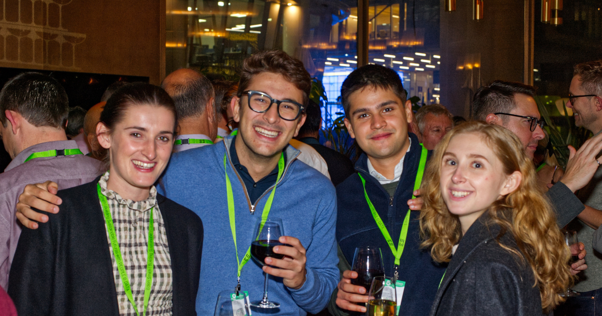 Four friends smiling at a nighttime event, holding drinks and wearing green lanyards.