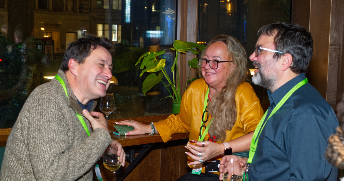 Three people in conversation at an indoor gathering, holding drinks and smiling near a table.