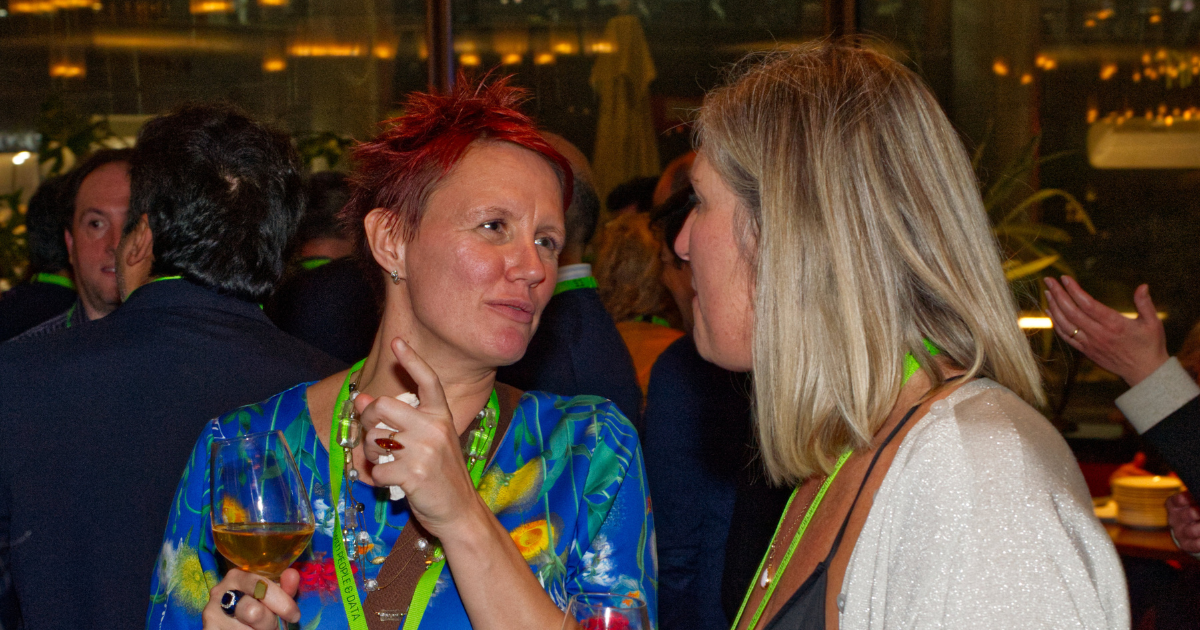 Two women talking at a crowded indoor event, one holding a drink, both wearing colorful outfits