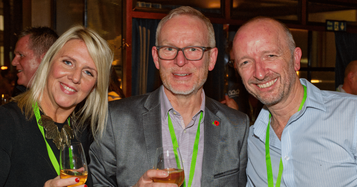 Three people smiling and holding drinks at a social event, wearing lanyards indoors.