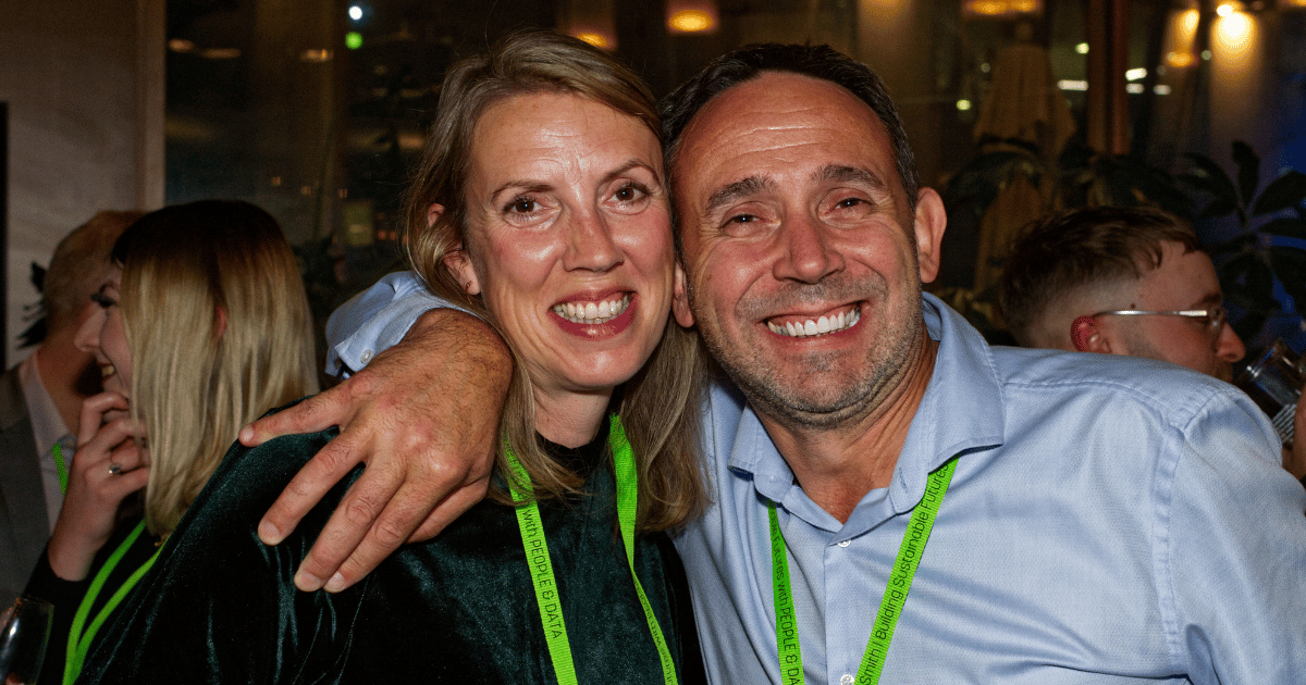Two smiling people pose closely together at a nighttime event, wearing lanyards.