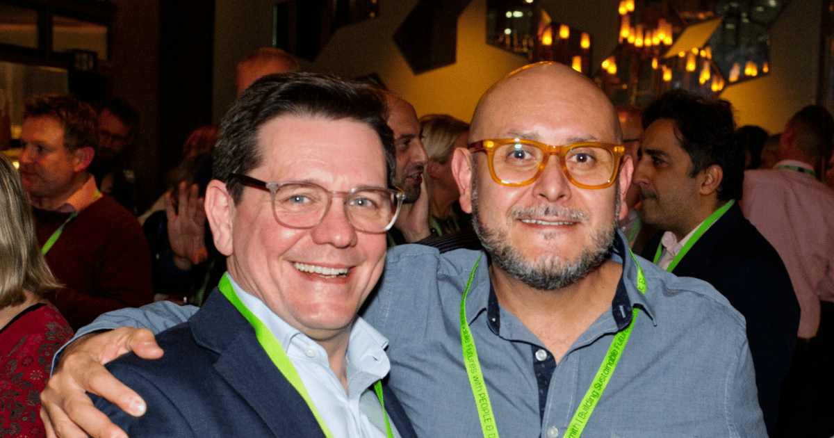 Two men smiling and posing together at a warmly lit indoor gathering