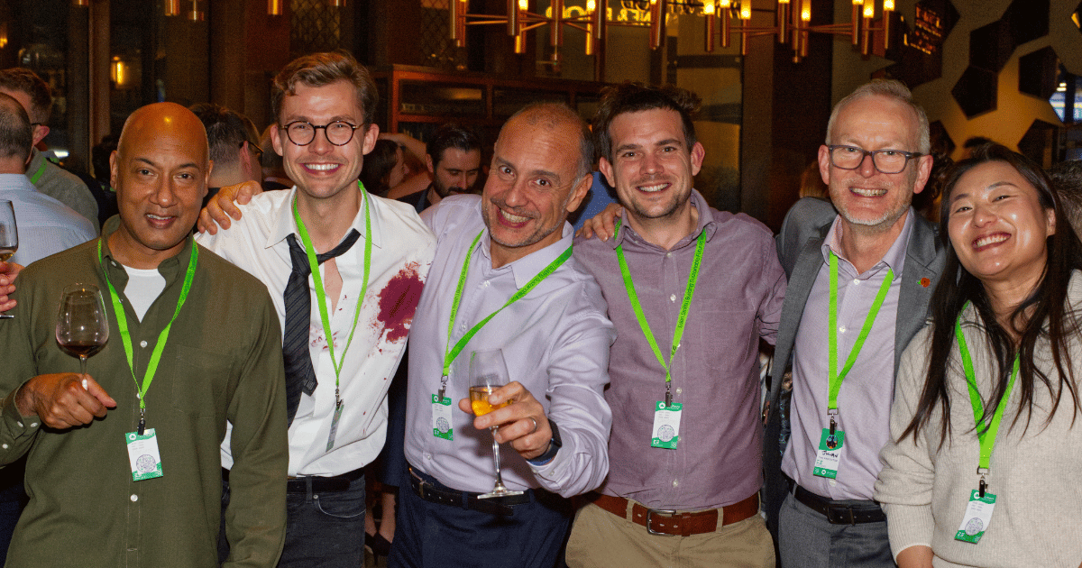 Six smiling people posing together at an indoor event, wearing green lanyards and holding drinks