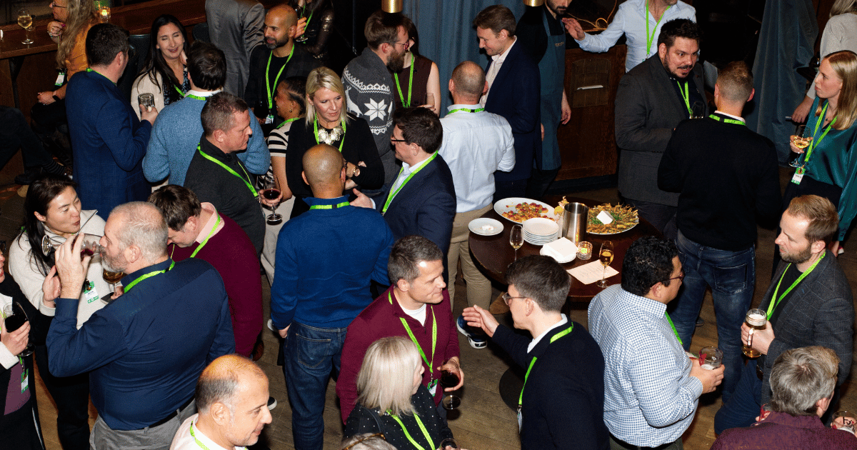 Crowded indoor gathering with people chatting and drinking, wearing green lanyards around buffet tables