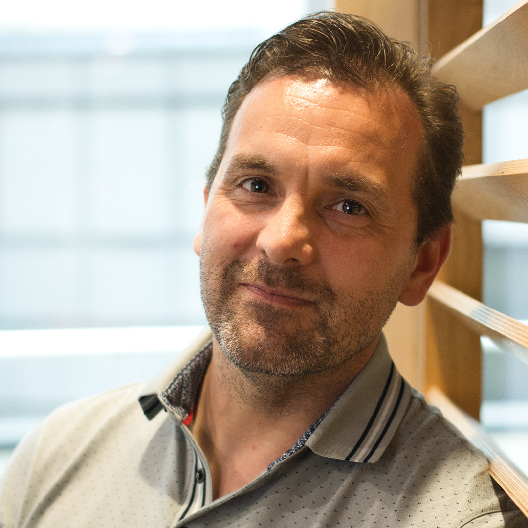 Man leaning against wooden railing, softly smiling in a bright indoor setting