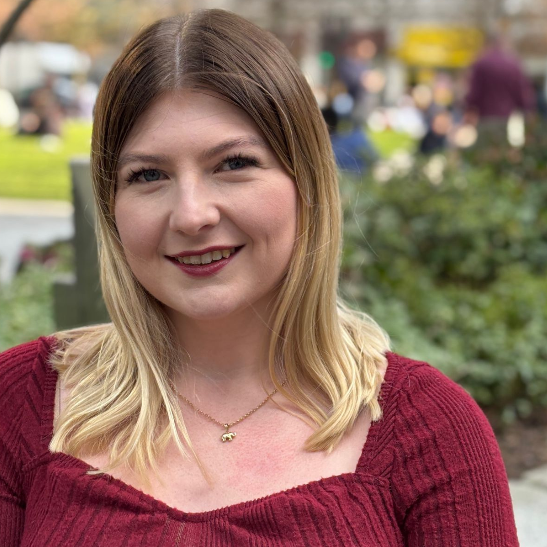 Portrait of a smiling woman in a burgundy top outdoors with blurred greenery behind her