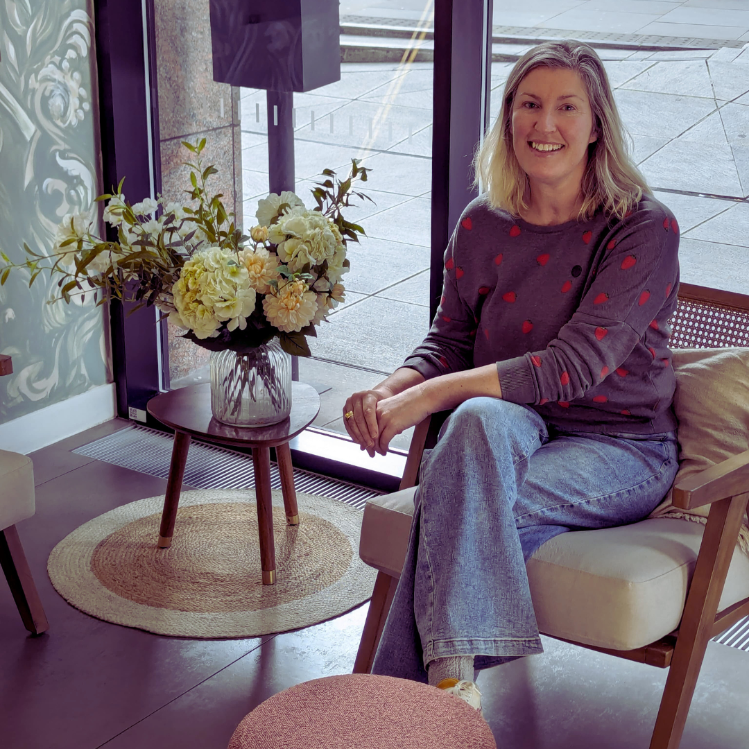 Woman seated by a window beside a vase of white flowers in a bright living room