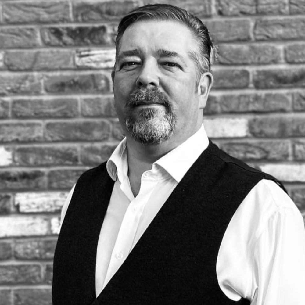 Portrait of a man in a white shirt and dark vest standing before a brick wall