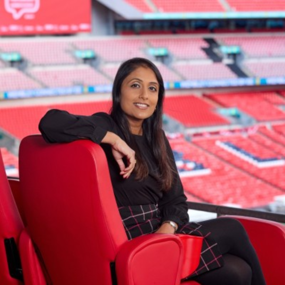 Person sitting in a red stadium seat, smiling with arms resting on the chair in a football stadium.