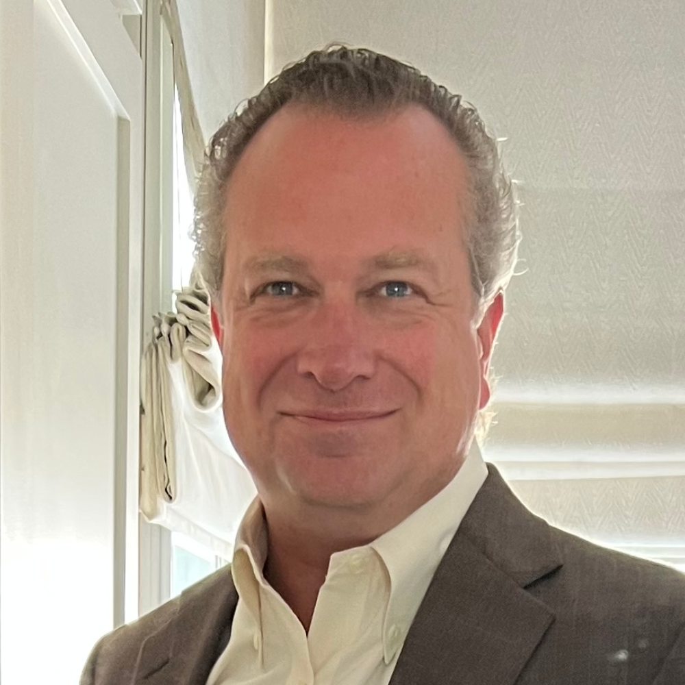 Smiling man in a light shirt and brown blazer standing indoors near a bright window.