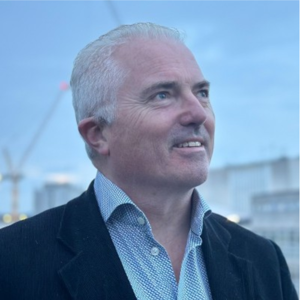 Smiling man in a dark coat and patterned shirt outdoors against a cloudy city skyline