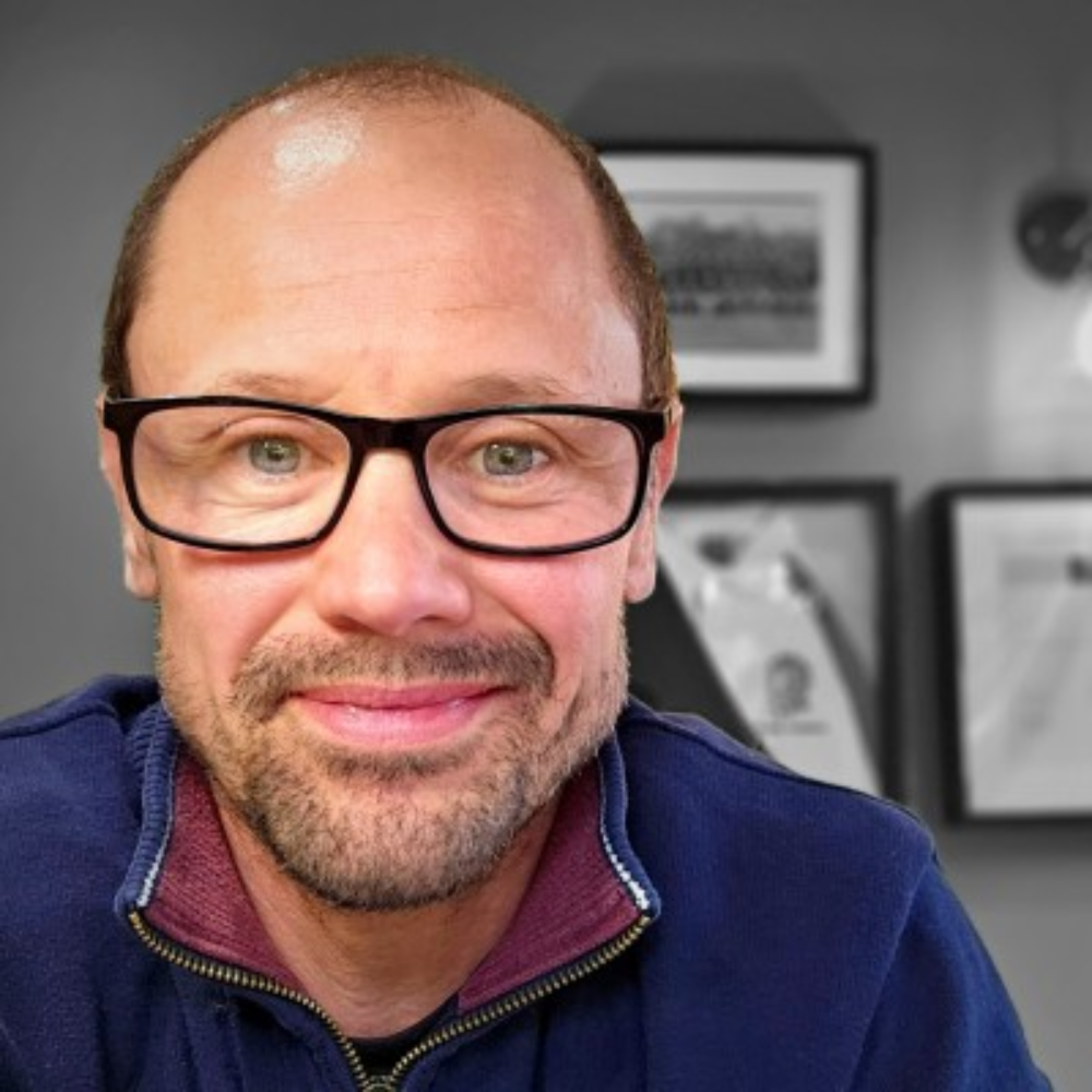 Smiling man with glasses in a blue jacket, sitting indoors with framed pictures behind him