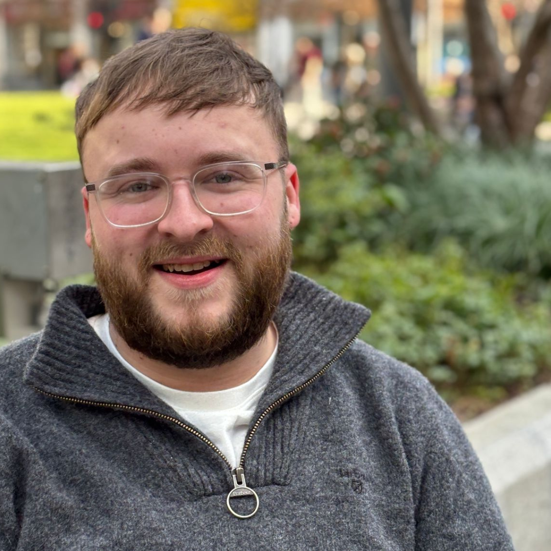 Smiling person with glasses and a beard outdoors in a gray sweater, with a blurred garden background