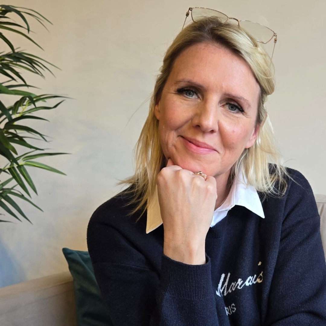 Blonde woman smiling with chin on hand, wearing a navy sweater, seated indoors beside a plant.