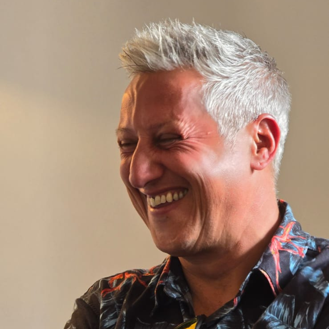Smiling person with short gray hair in a patterned shirt, looking down against a plain background.