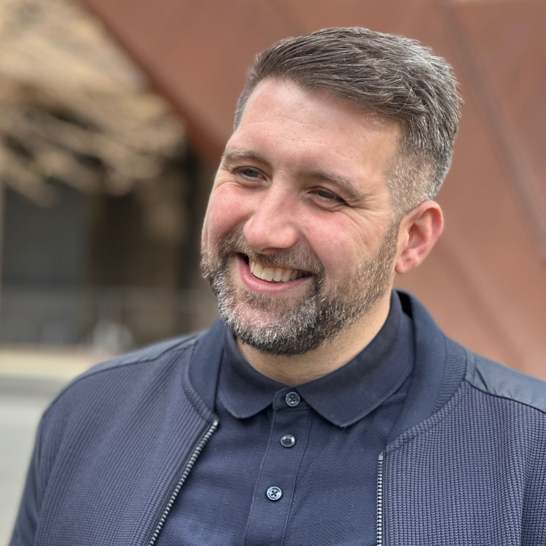 Smiling man in a navy jacket outdoors, with a blurred building in the background