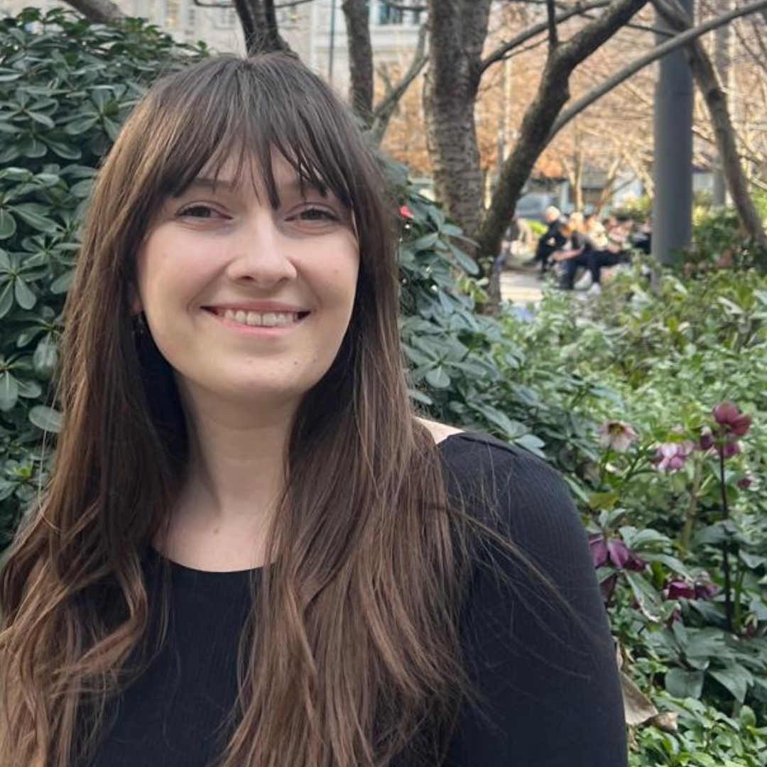 Smiling person with long brown hair in front of leafy shrubs and trees outdoors