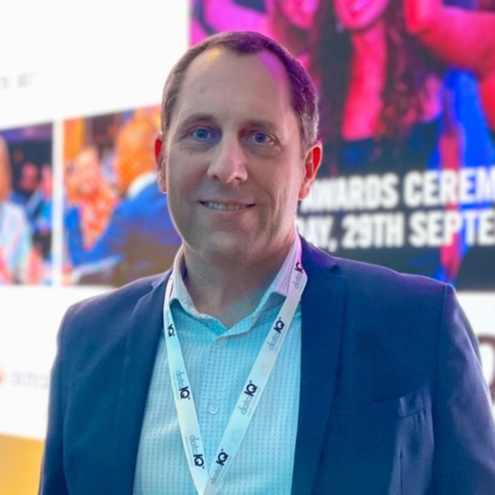 Man in a blue suit and lanyard smiling at a conference with colorful event signage behind him