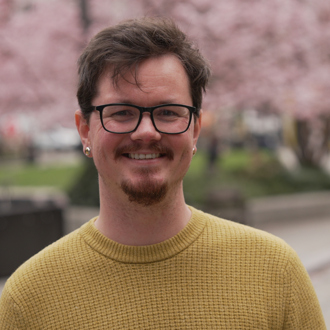 Smiling person with glasses and a mustard sweater, standing outdoors with pink blossoms blurred behind.